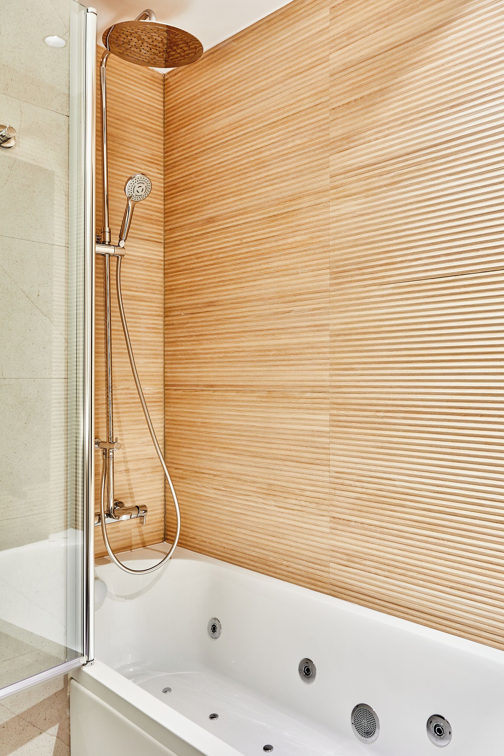 A modern bathroom with a glass-enclosed shower, a wooden wall, and a white bathtub with multiple faucets.