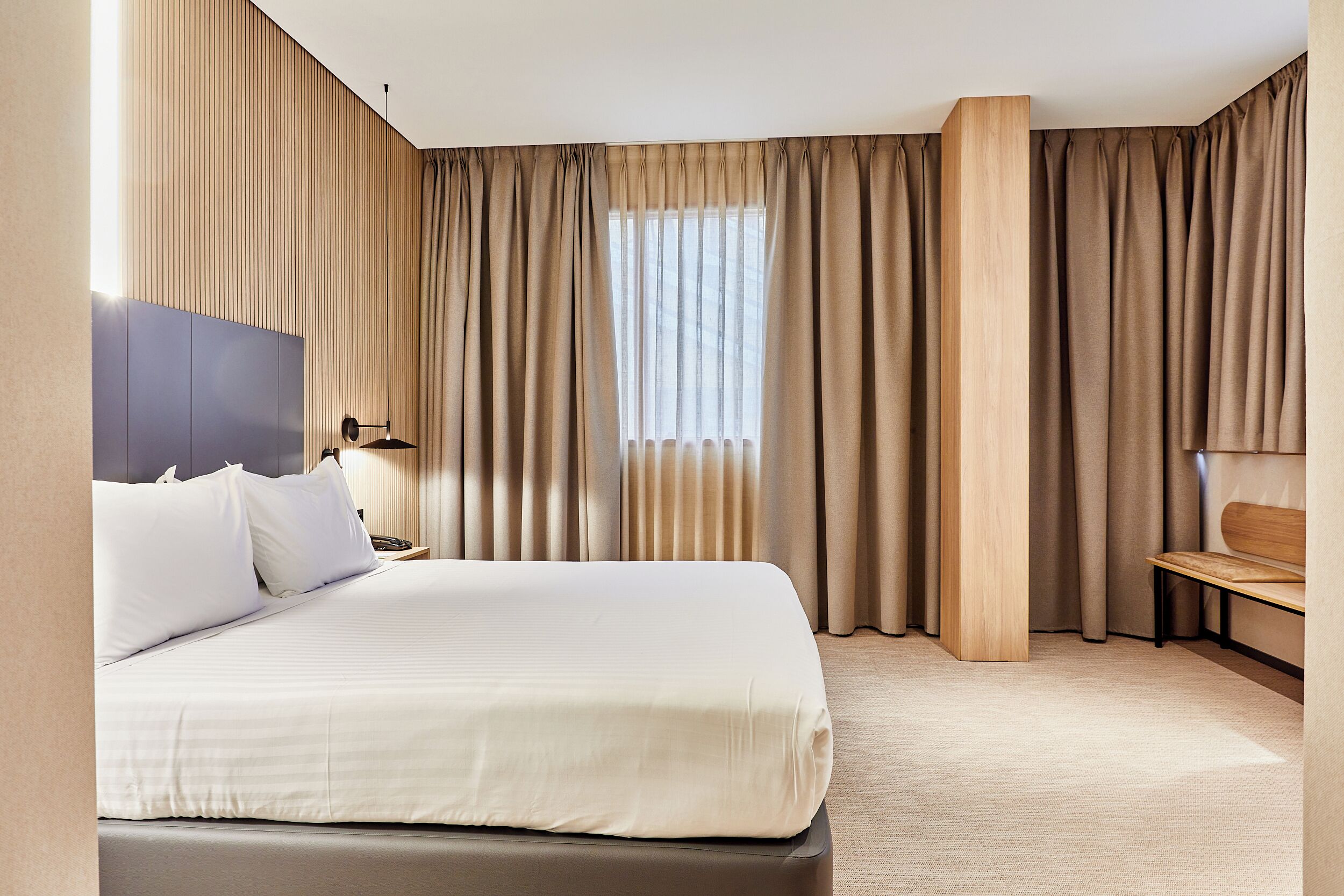 Une chambre d’hôtel avec un grand lit, une tête de lit en bois, une table de chevet, un bureau et une fenêtre avec des rideaux.