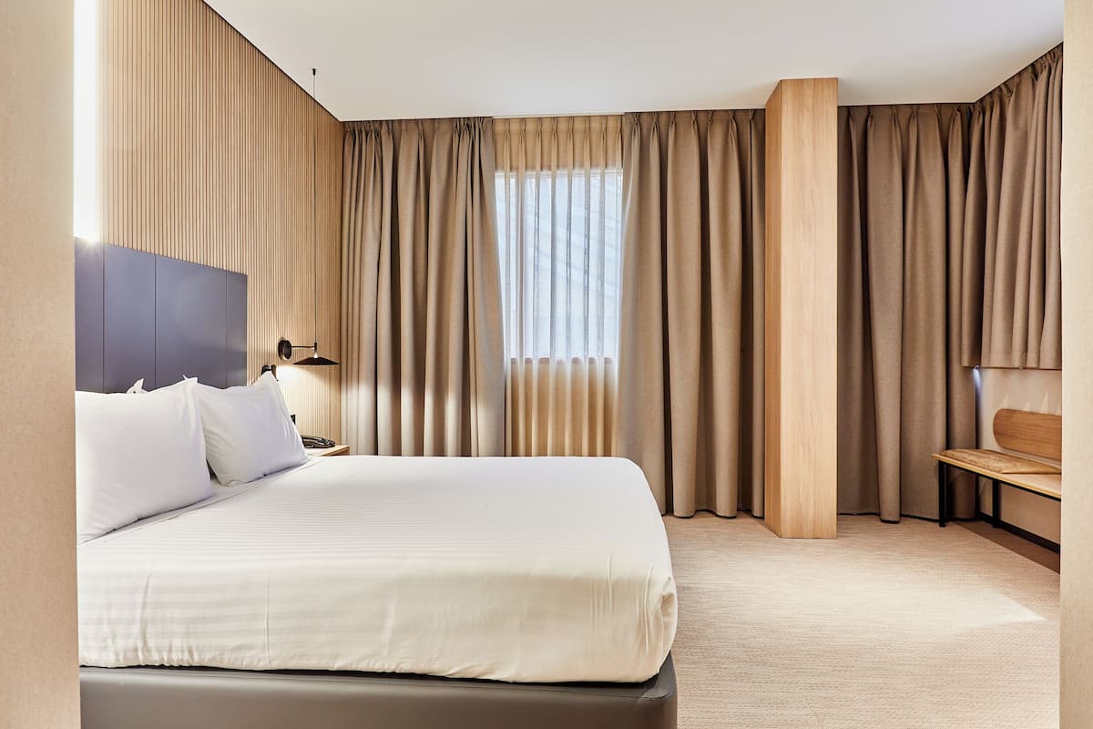 A hotel room with a large bed, a wooden headboard, a nightstand, a desk, and a window with curtains.