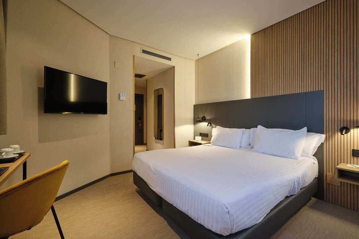 Ein modernes Hotelzimmer mit einem großen Bett, einem flachbildfernseher an der Wand, einem Schreibtisch mit Stuhl und einer Wandverkleidung aus Holz.