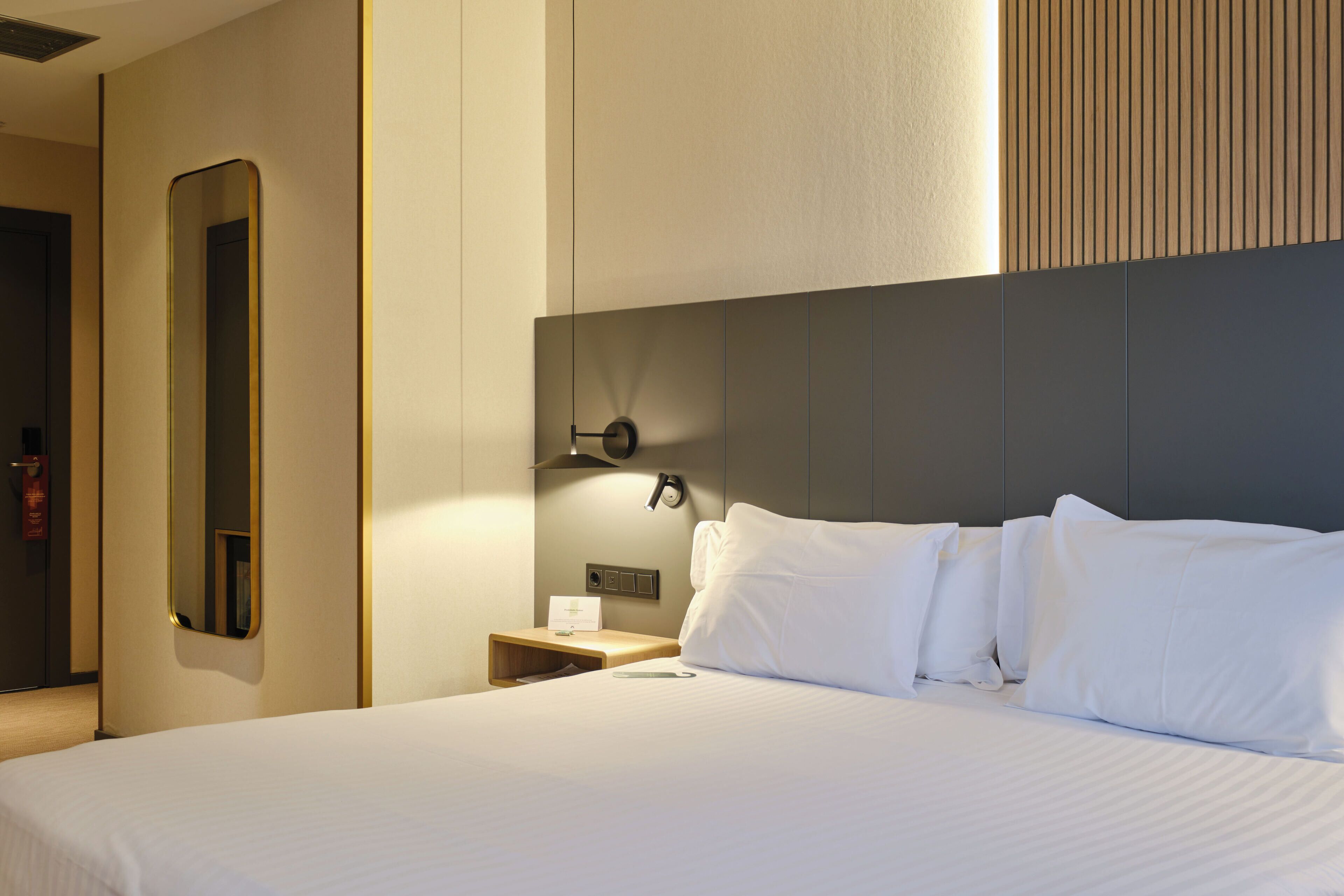 A hotel room with a large bed, a nightstand, a mirror, and a wall-mounted light fixture.