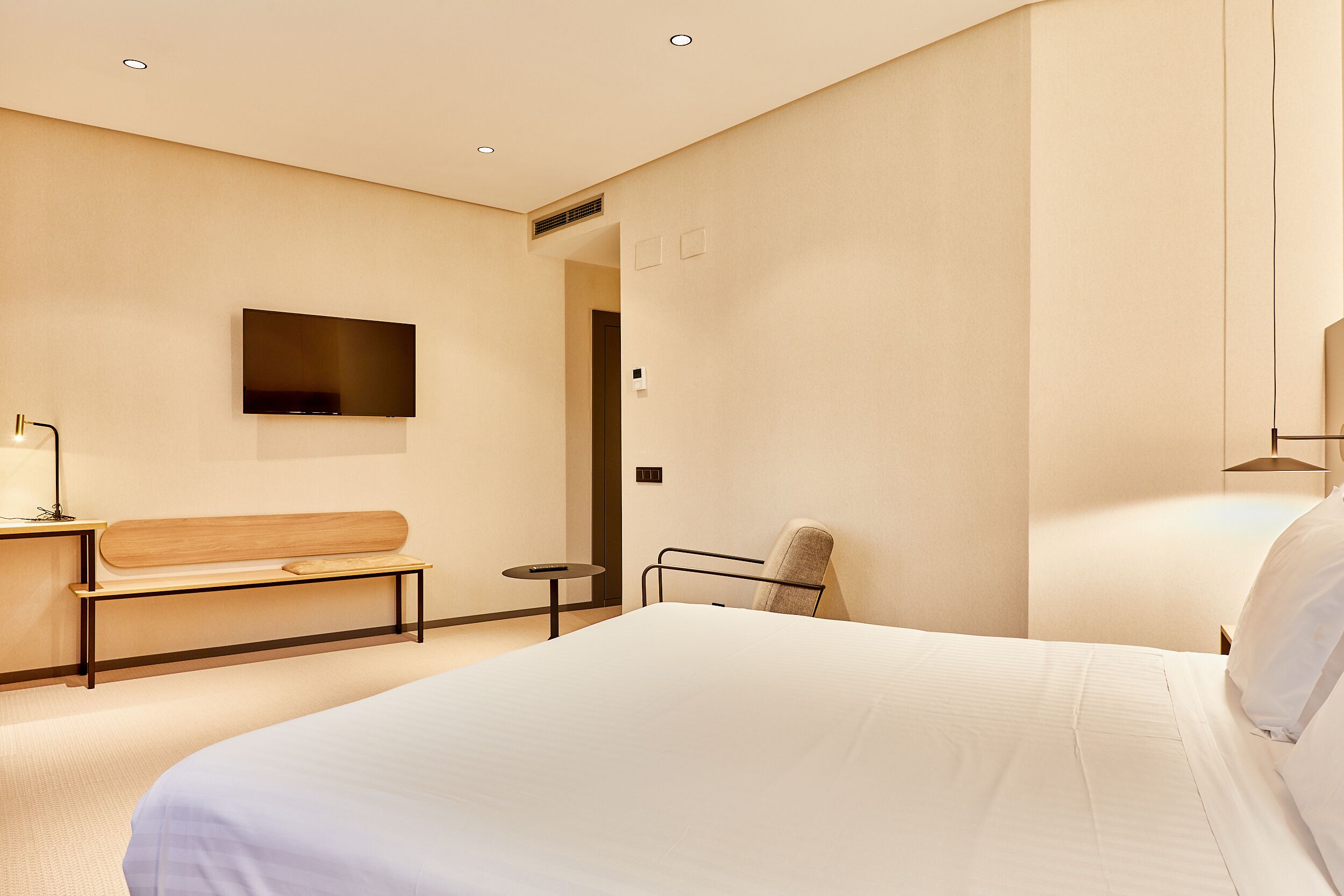 A modern hotel room with a large bed, a flat-screen TV mounted on the wall, a bench, and a small table.