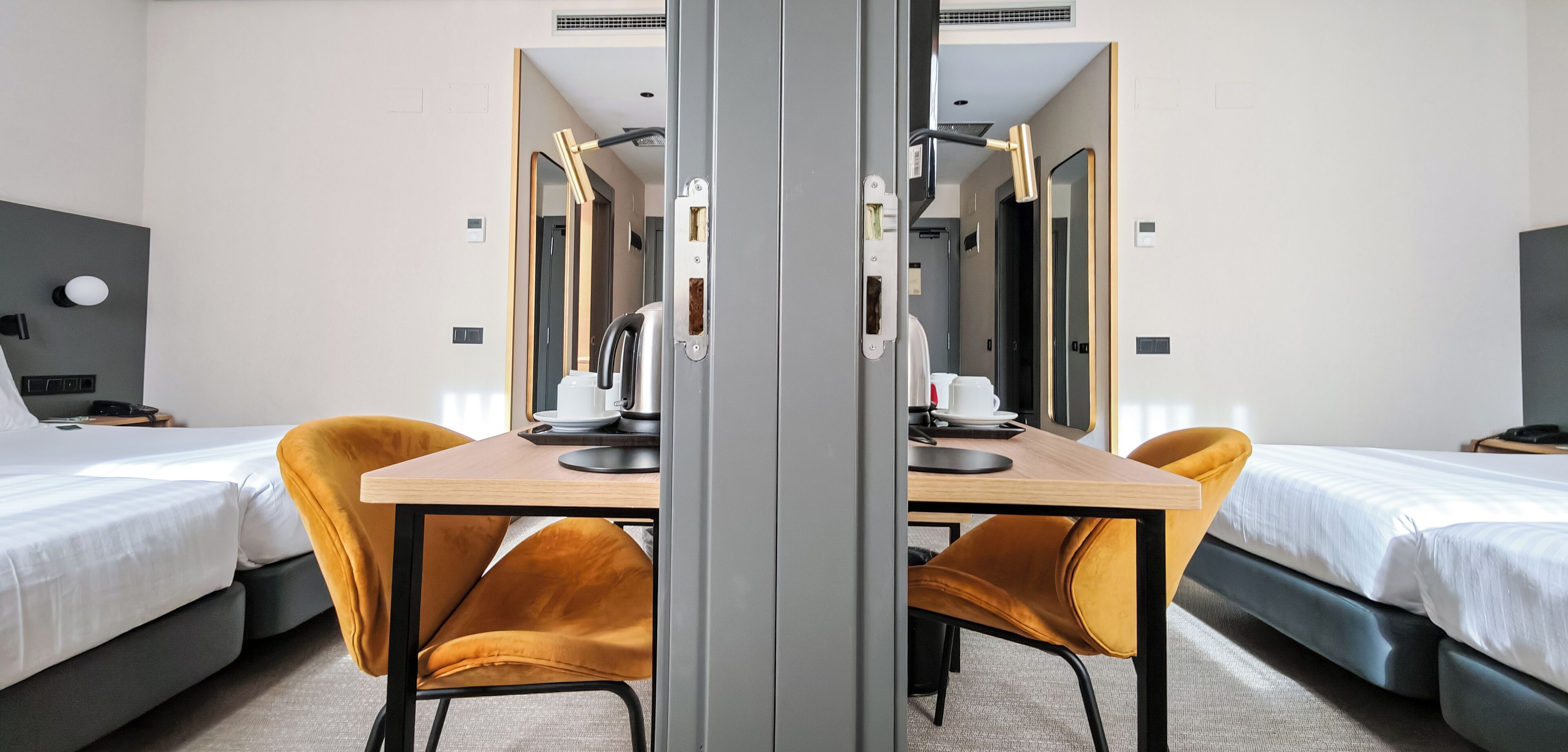 A hotel room with two beds, a wooden table with chairs, a mirror, and a desk with a computer and a cup.
