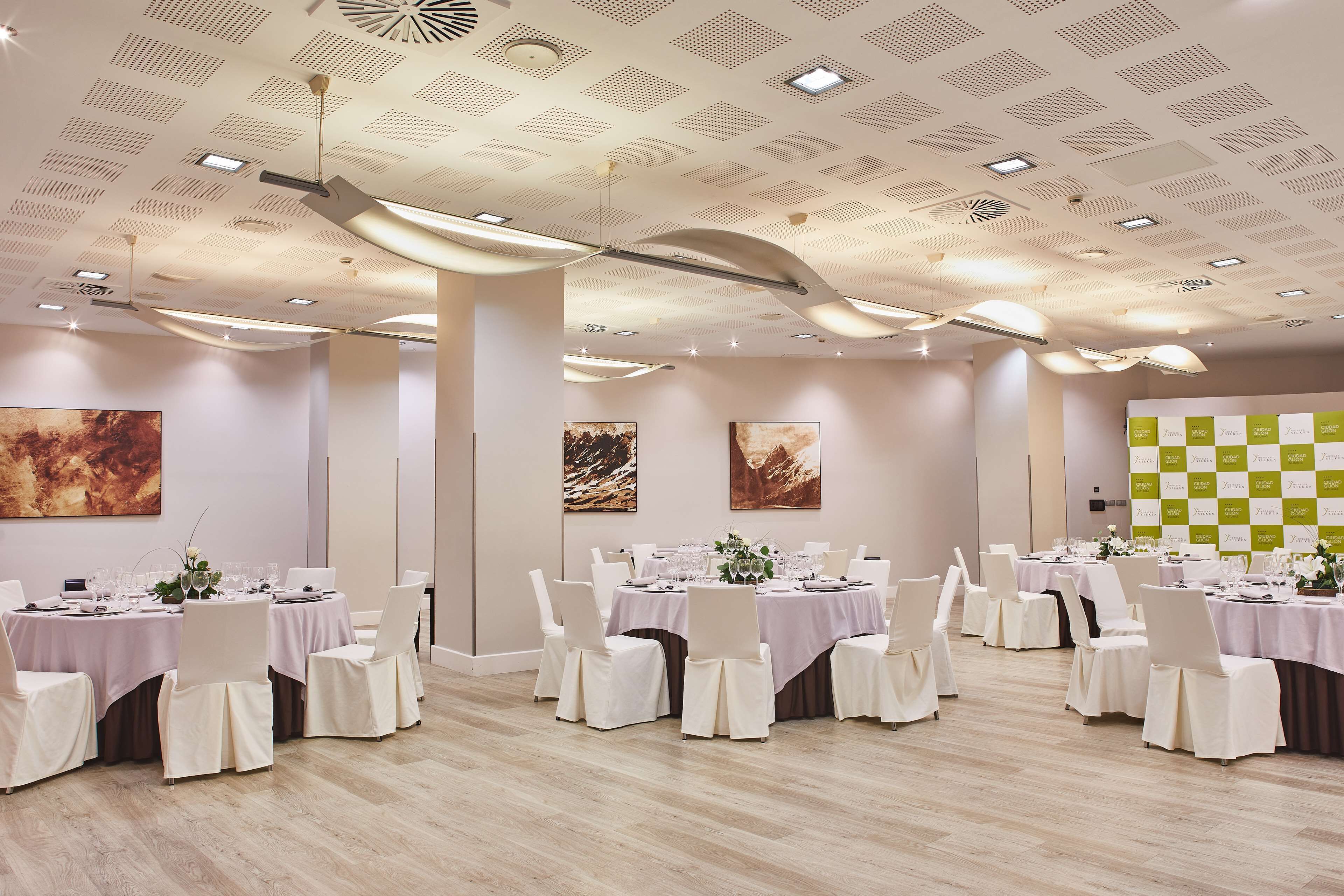 Un amplio salón de banquetes con mesas redondas, sillas blancas y un diseño moderno en el techo.