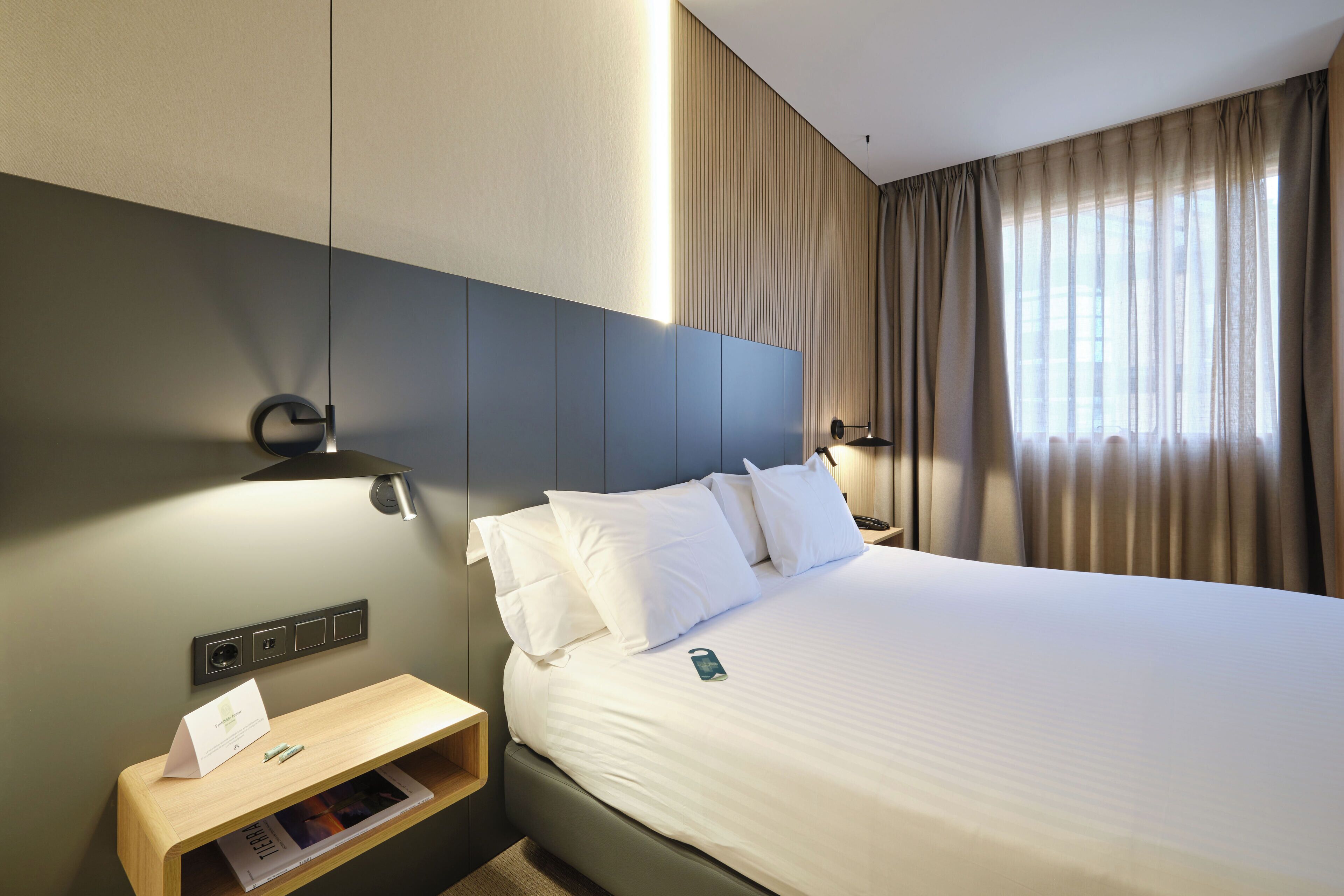 Una habitación de hotel con cama, mesita de noche, lámpara y ventana con cortinas.