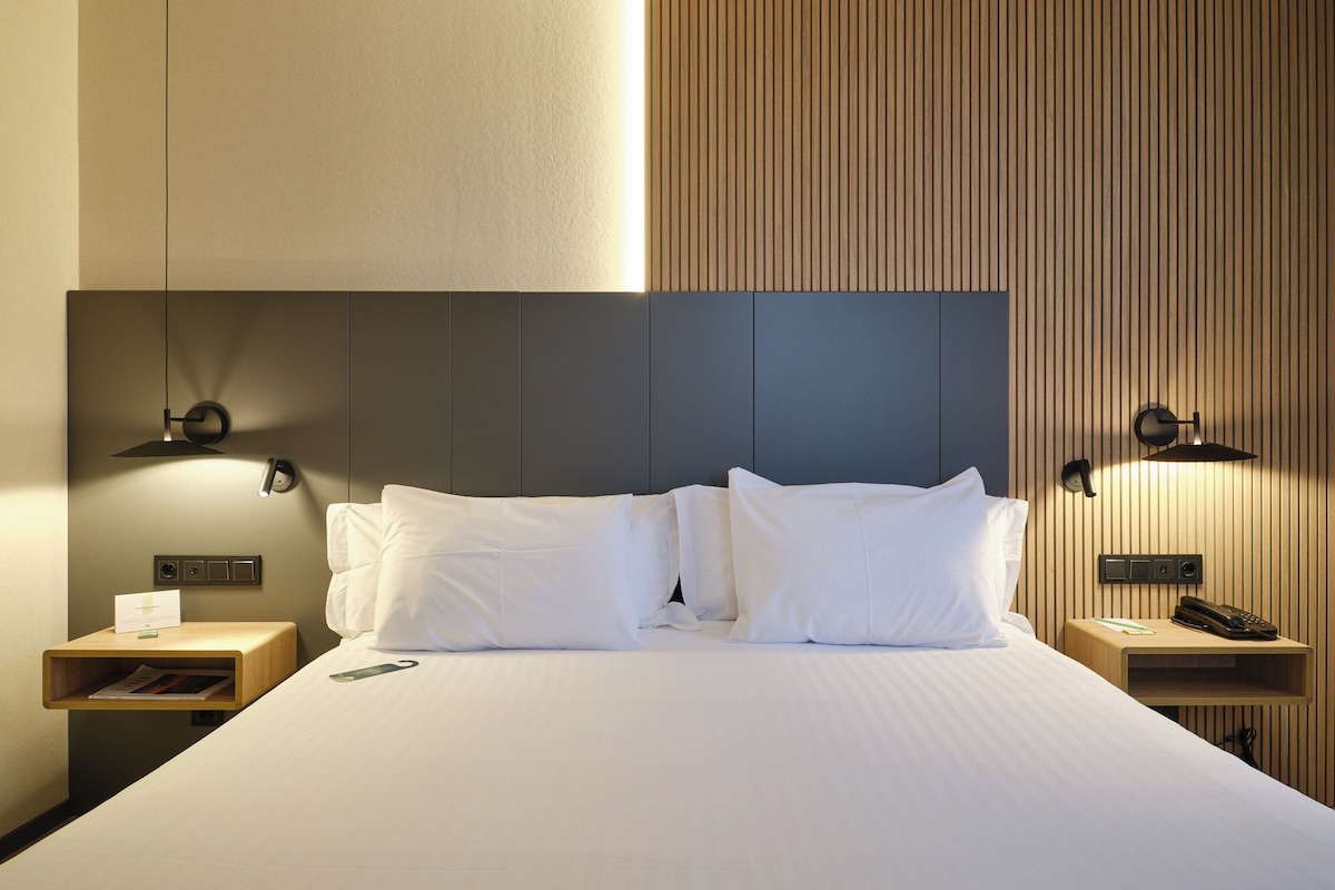 Una cama bien hecha con sábanas blancas, dos mesitas de noche con lámparas y un panel de pared de madera.