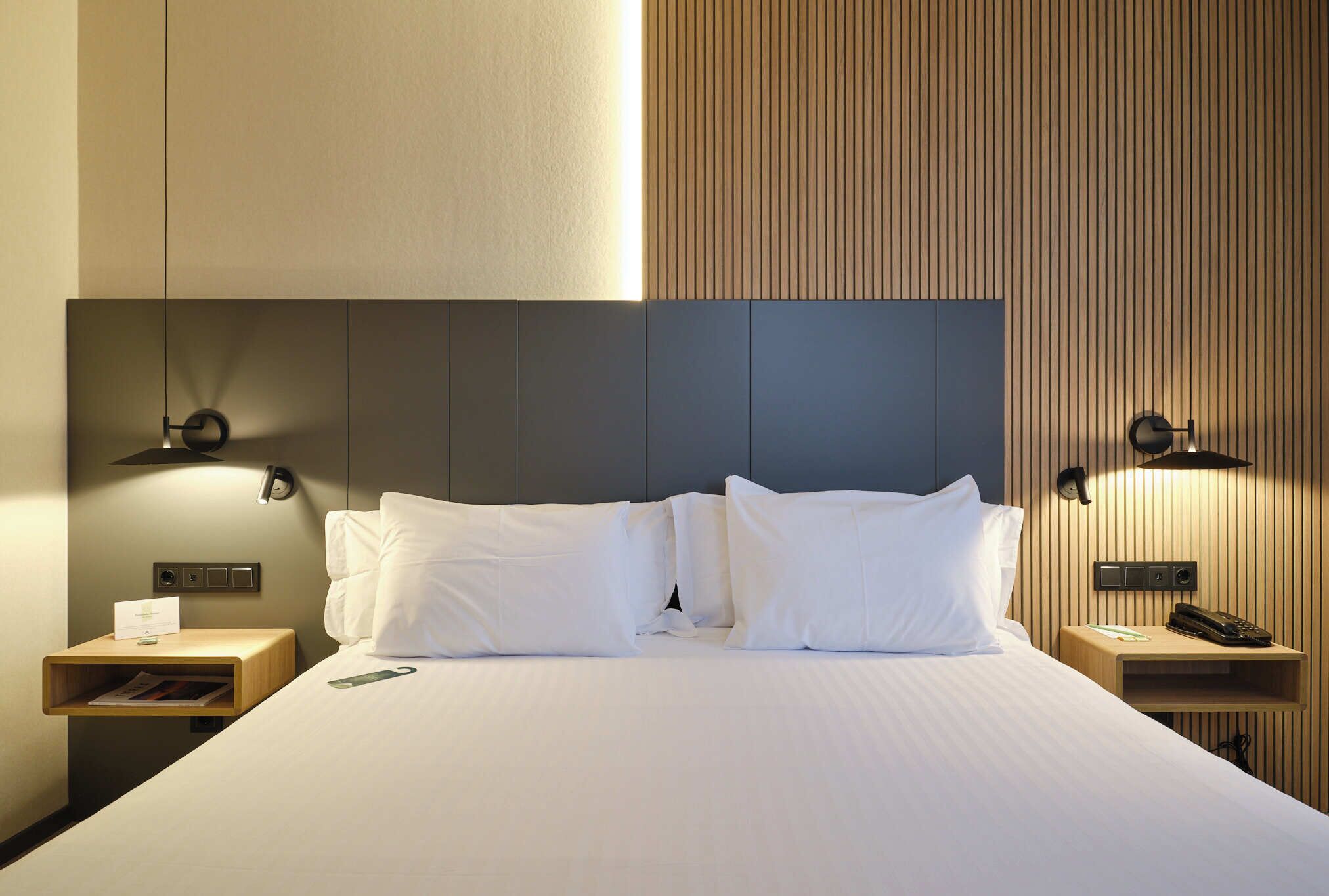 Una cama bien hecha con sábanas blancas, dos mesitas de noche con lámparas y un panel de pared de madera.