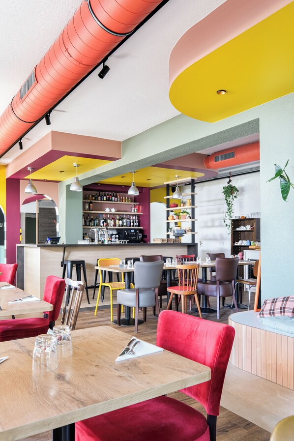 A vibrant restaurant interior with wooden tables, red and yellow chairs, and a bar area with shelves of bottles and a coffee machine.