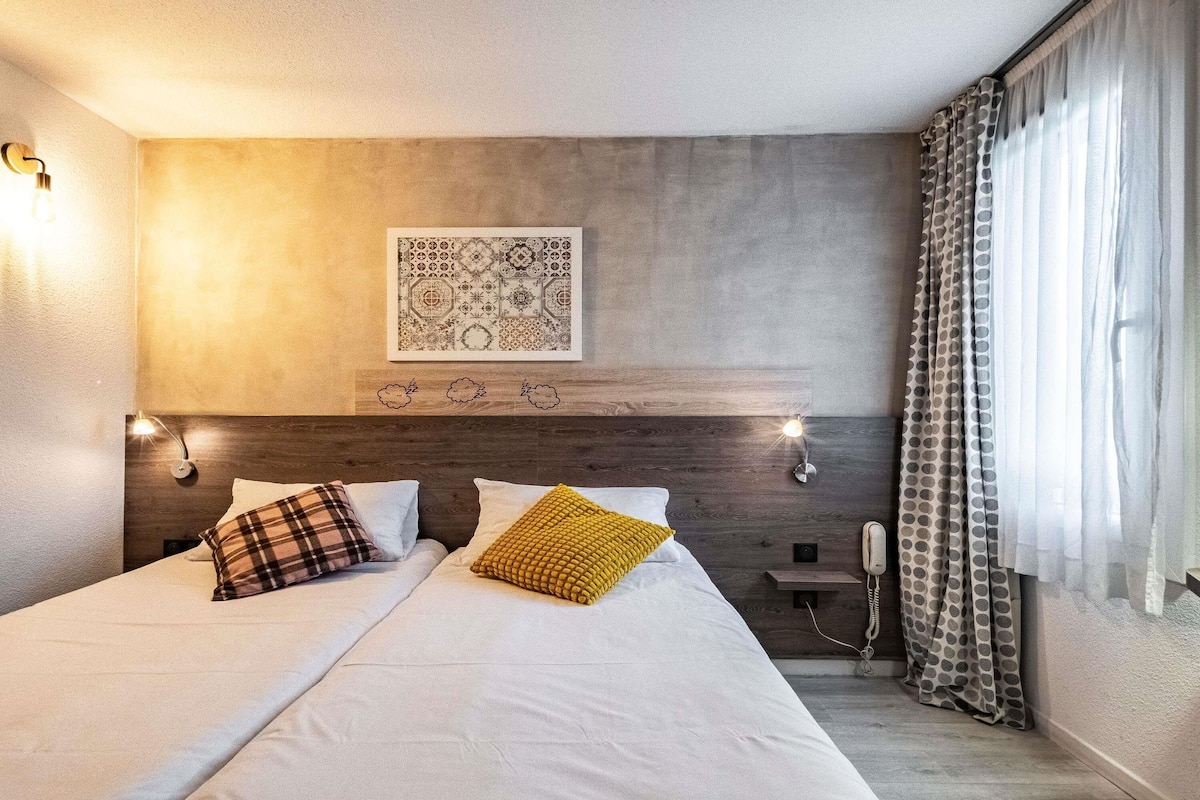 Quarto de hotel com cama, duas luminárias de cabeceira, telefone fixado na parede e janela com cortinas.