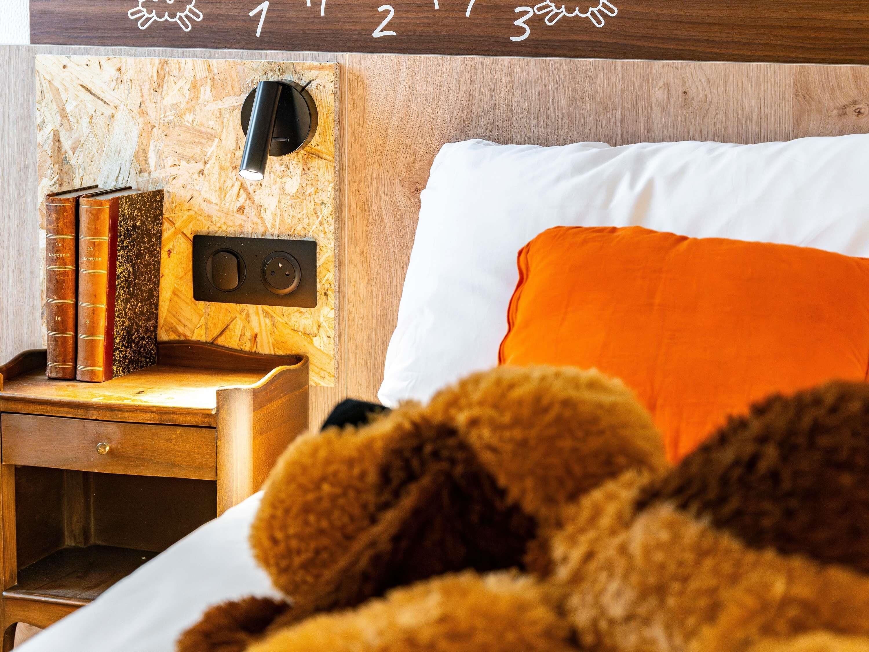 Un lit bien fait, agrémenté d’un coussin orange, une table de chevet en bois avec des livres, et un luminaire fixé au mur.