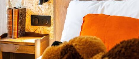 Un lit bien fait, agrémenté d’un coussin orange, une table de chevet en bois avec des livres, et un luminaire fixé au mur.