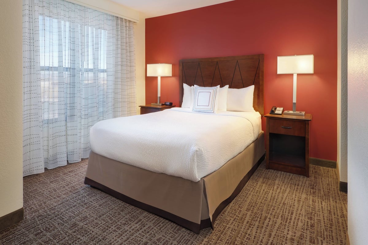 A hotel room with a large bed, two bedside tables with lamps, a nightstand, and a window with sheer curtains.