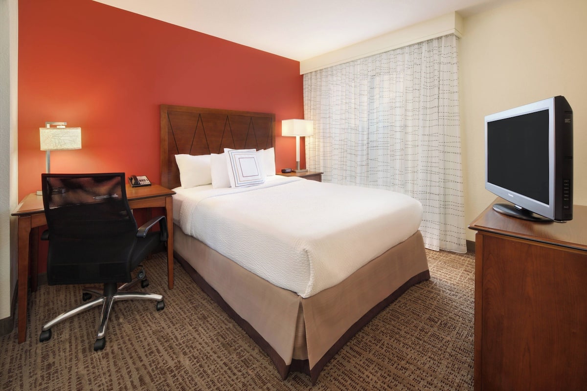 A hotel room with a large bed, two bedside tables, a desk with a chair, a television, and a window with curtains.