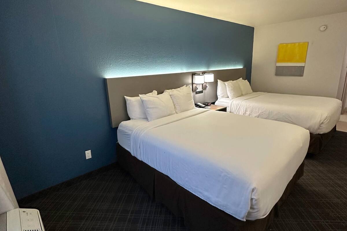 Hotellihuone, jossa on kaksi sänkyä, sängynpääty, yöpöytä lampun kanssa ja seinälle kiinnitetty taideteos.