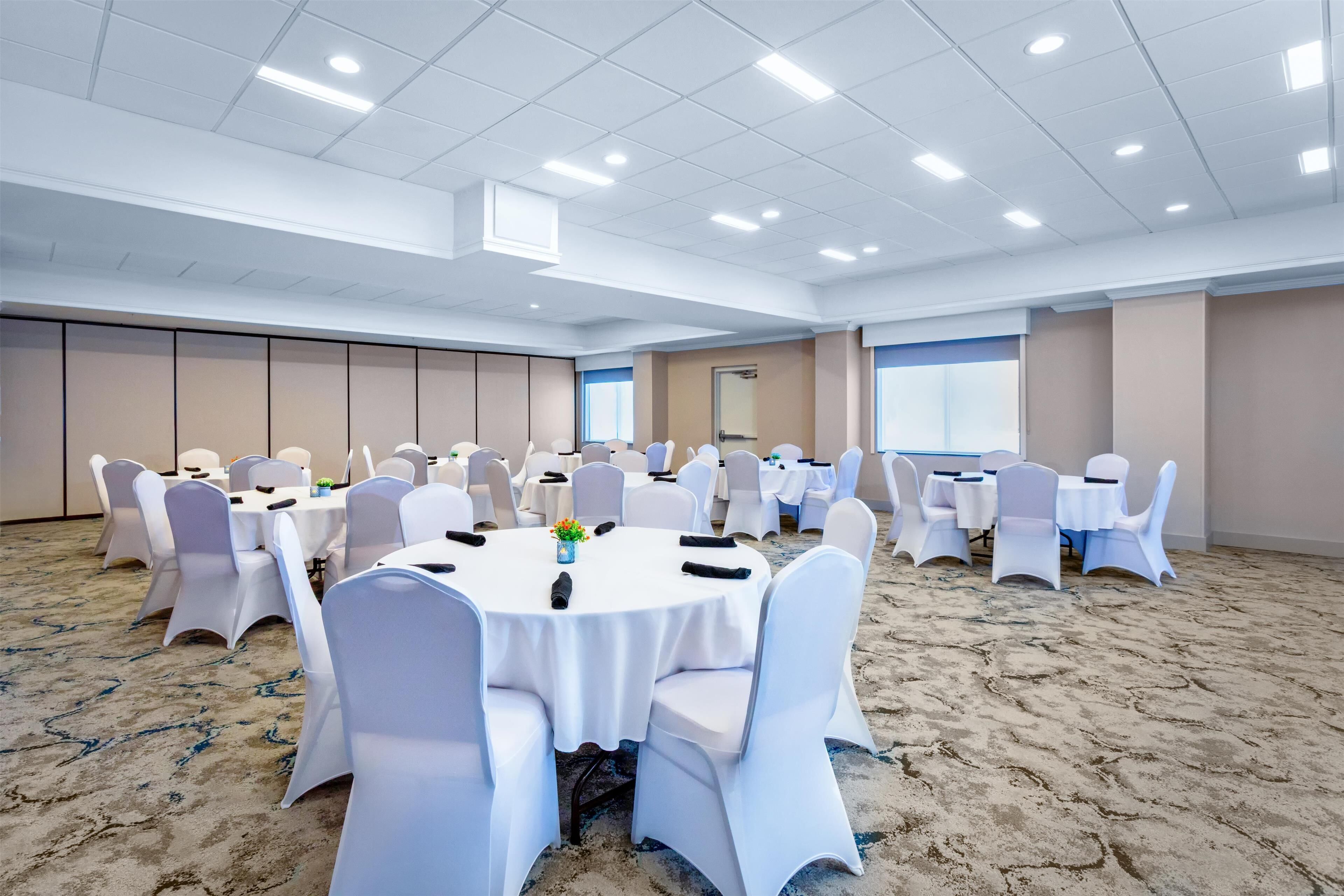 Un salón de conferencias con mesas redondas y sillas blancas, suelo alfombrado y un techo alto con iluminación empotrada.