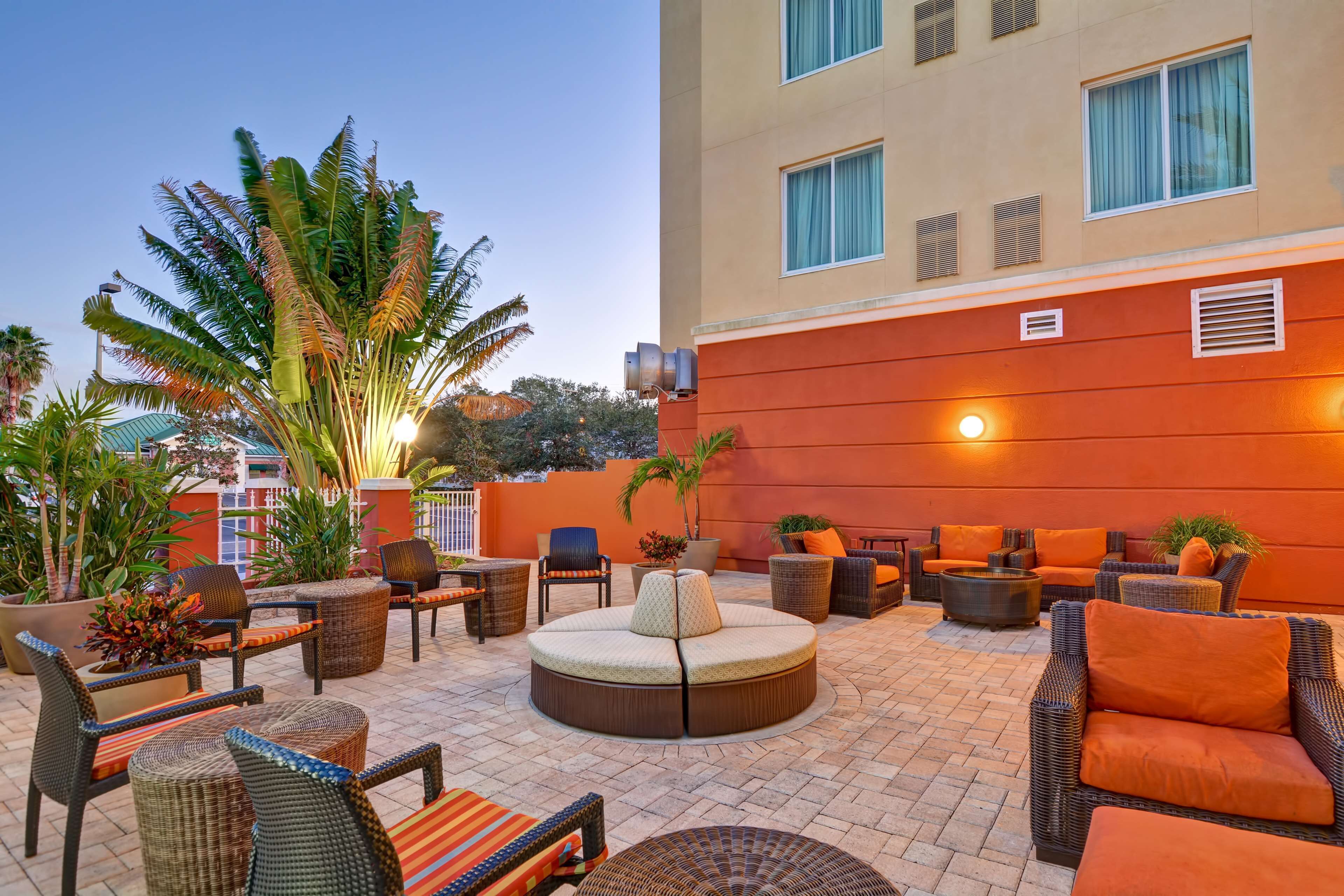 An outdoor seating area with wicker furniture and a round table, surrounded by potted plants and a building with large windows.
