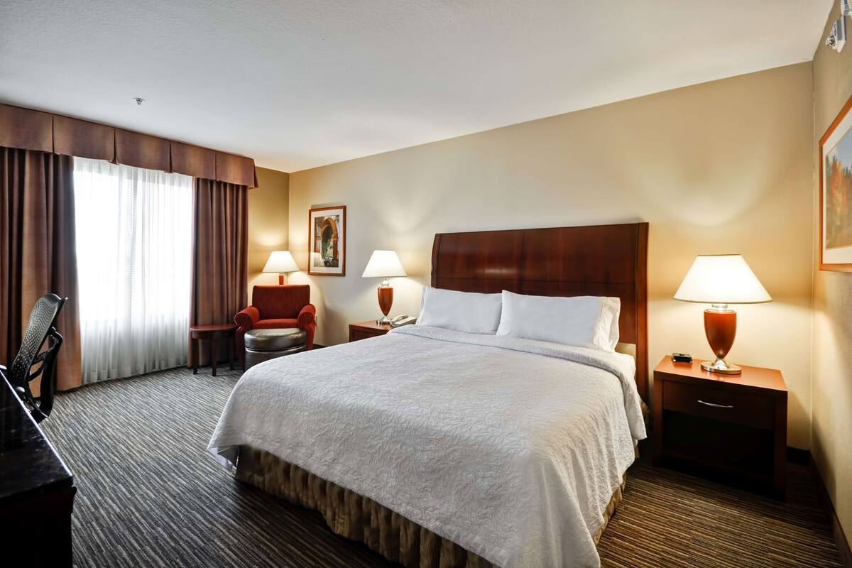 A hotel room with a large bed, two bedside tables with lamps, a chair, a desk, and a window with curtains.