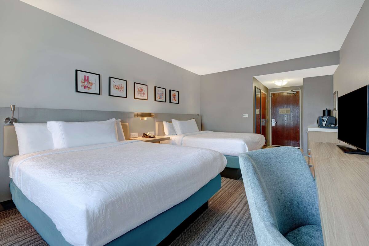 A hotel room with two beds, a desk, a chair, a TV, and framed artwork on the wall.