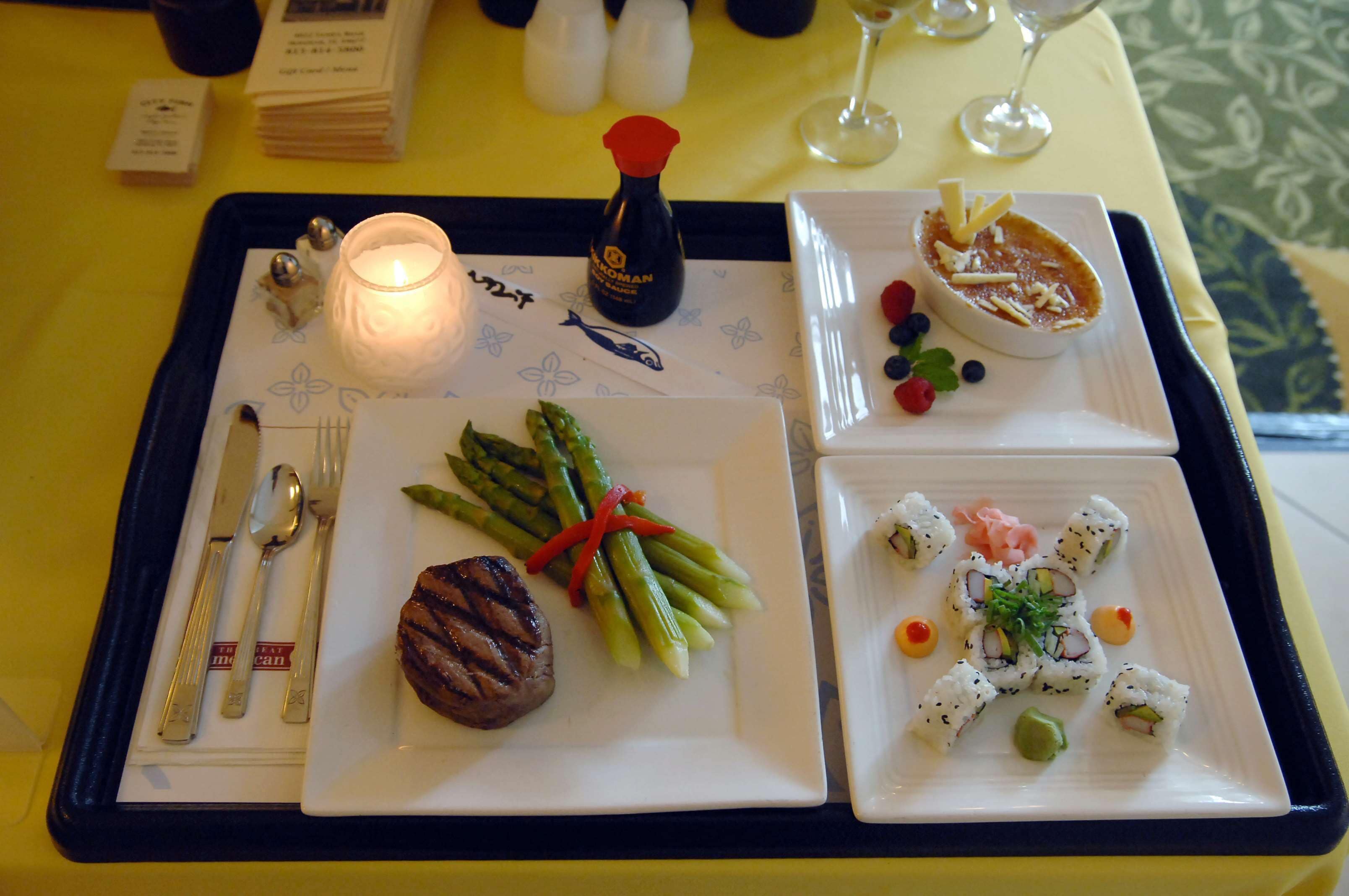 A tray with a grilled steak, asparagus, and sushi on a table with a candle and a bottle of sauce.