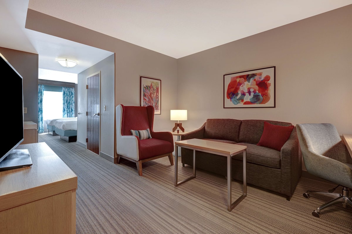 A hotel room with a bed, a desk with a laptop, a red armchair, a brown sofa, a small table with a lamp, and two abstract paintings on the walls.