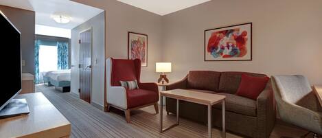 A hotel room with a bed, a desk with a laptop, a red armchair, a brown sofa, a small table with a lamp, and two abstract paintings on the walls.