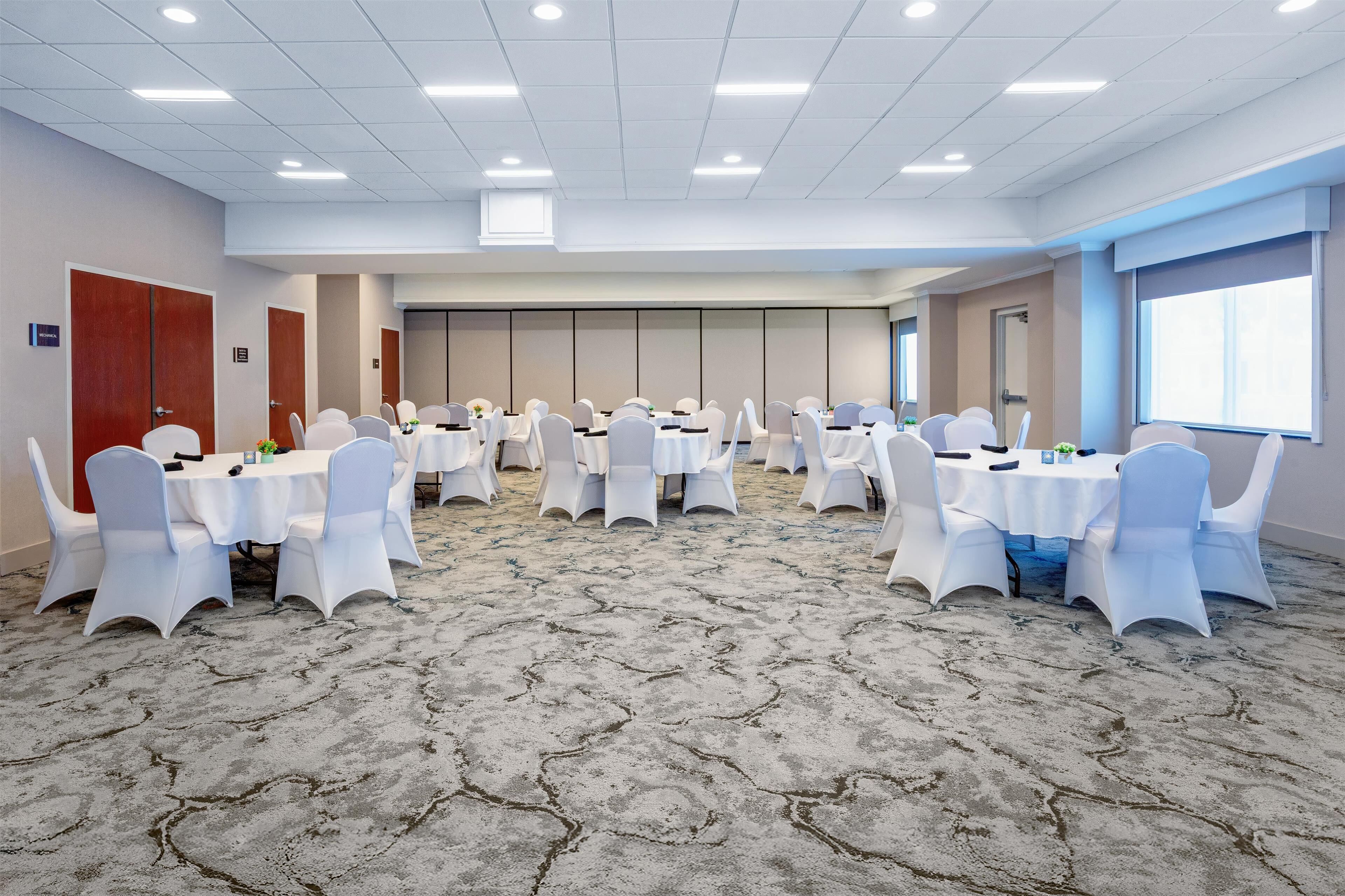 A conference room with multiple tables and chairs, carpeted floor, and a large window.