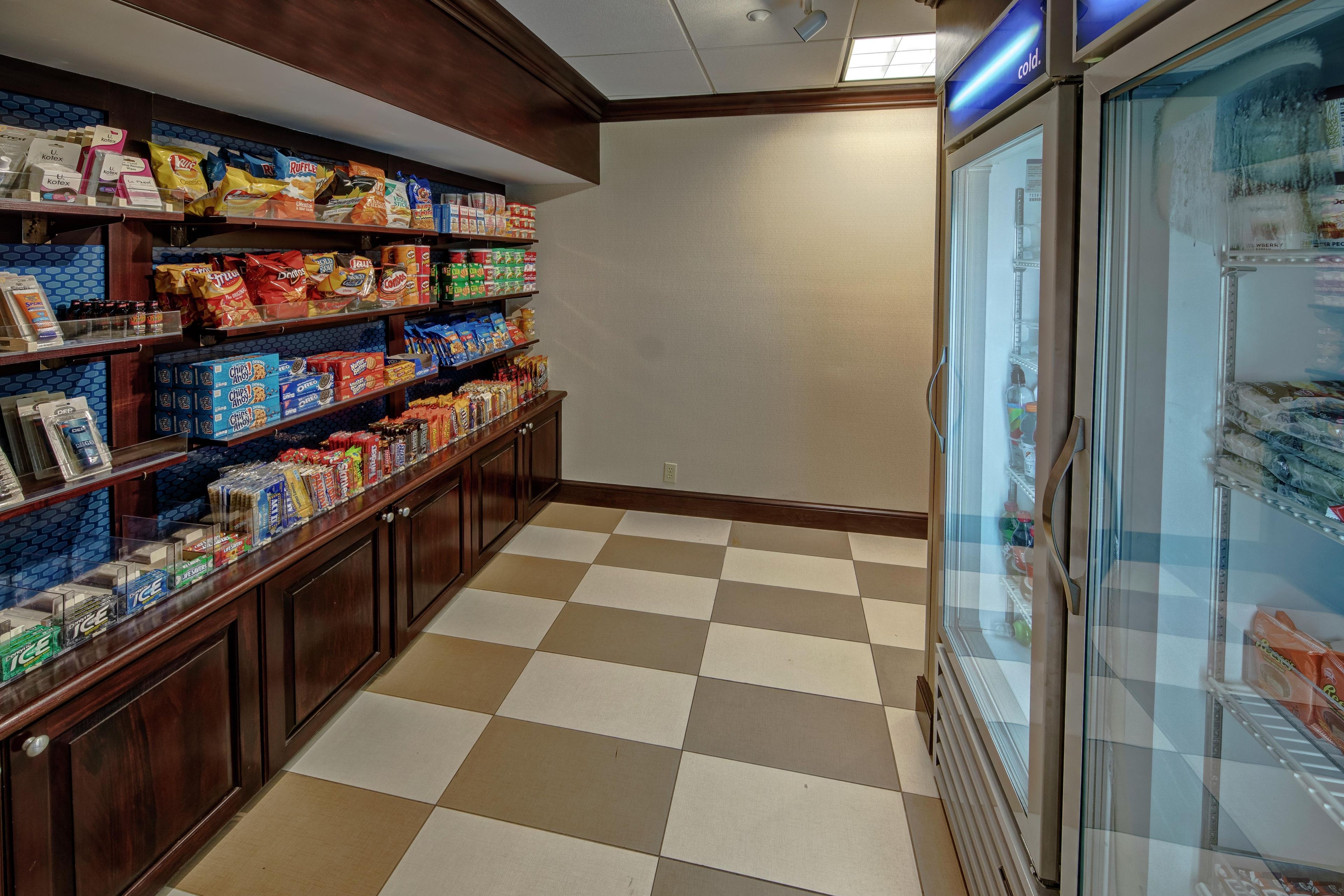 A grocery store interior with shelves stocked with snacks and beverages, a checkered floor, and a refrigerated display case.