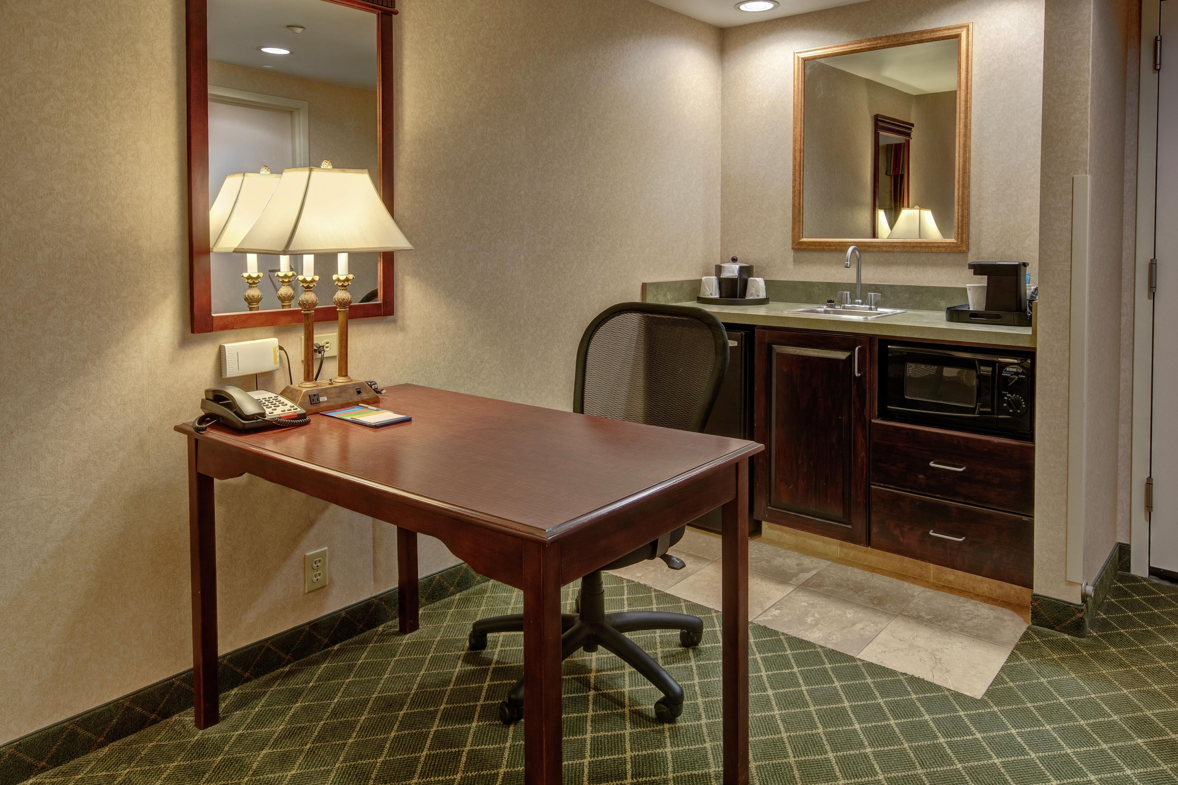 A hotel room with a wooden desk, a telephone, a lamp, a chair, a sink, a mirror, and a microwave.