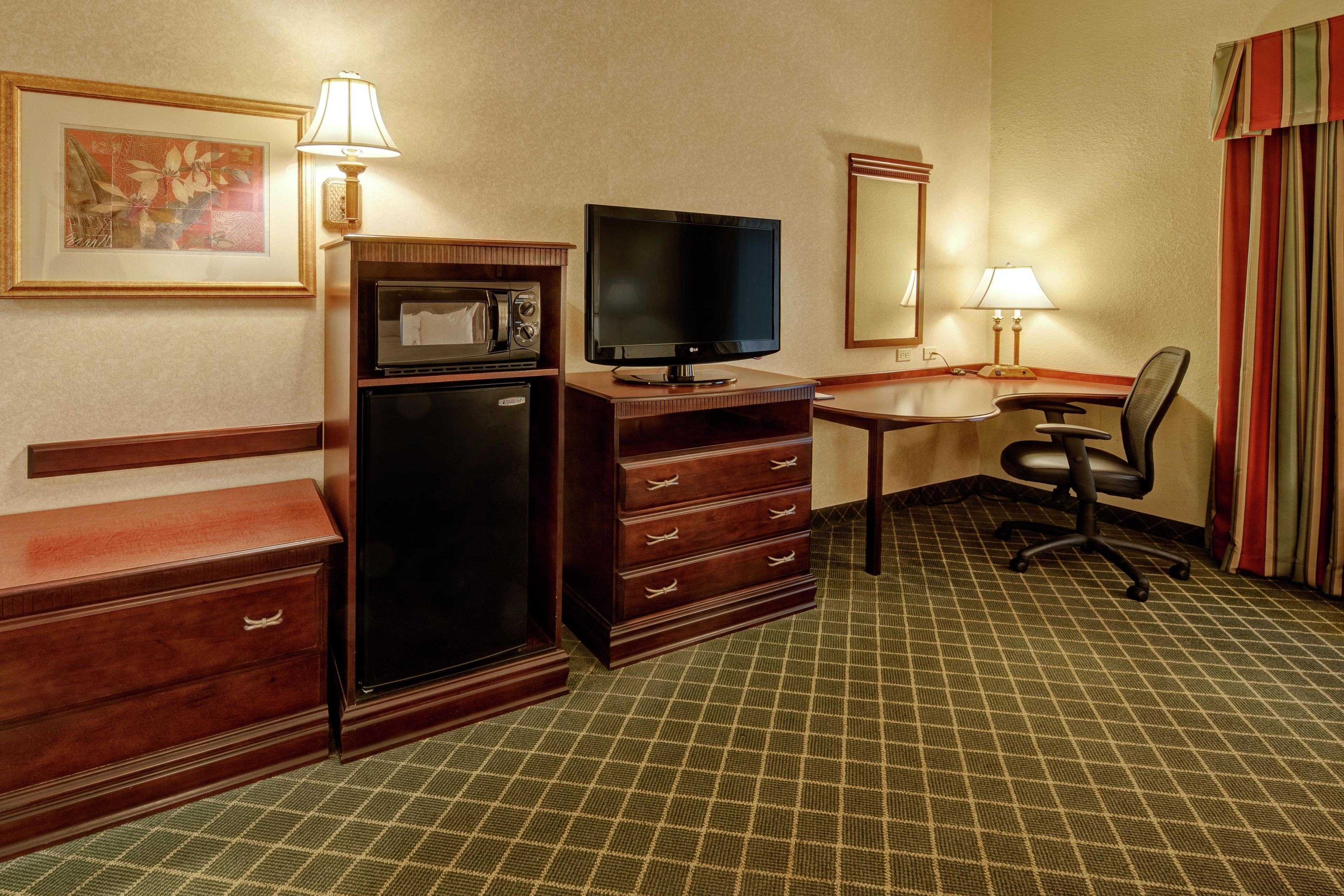 A hotel room with a kitchenette, a flat-screen TV, a desk with a chair, and a patterned carpet.