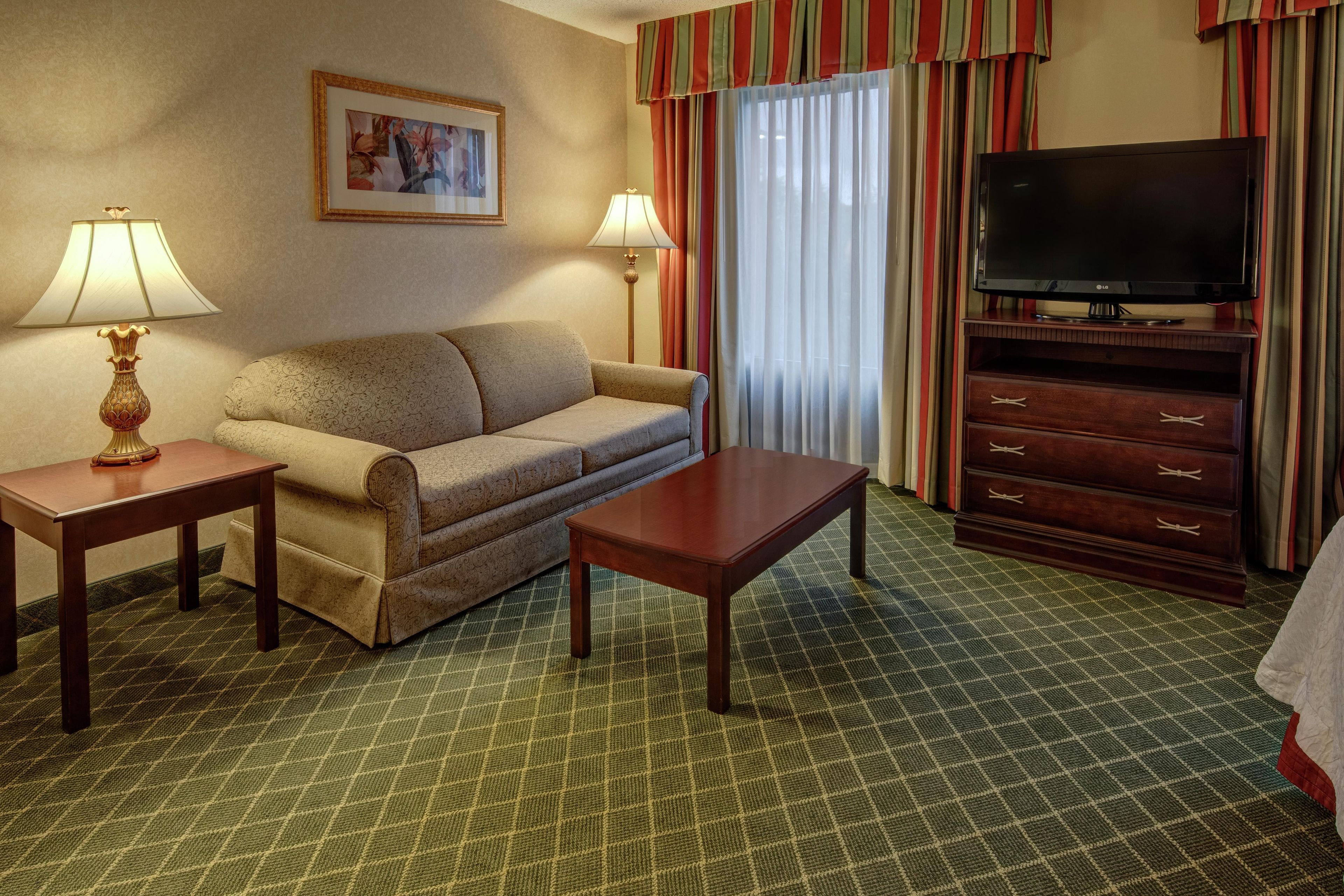 A hotel room with a sofa, a coffee table, a TV on a stand, two lamps, a painting on the wall, and a patterned carpet.