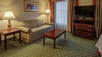 Habitación de hotel con sofá, una mesa de centro, televisor sobre un mueble, dos lámparas, un cuadro en la pared y alfombra estampada.