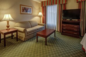 Habitación de hotel con sofá, una mesa de centro, televisor sobre un mueble, dos lámparas, un cuadro en la pared y alfombra estampada.
