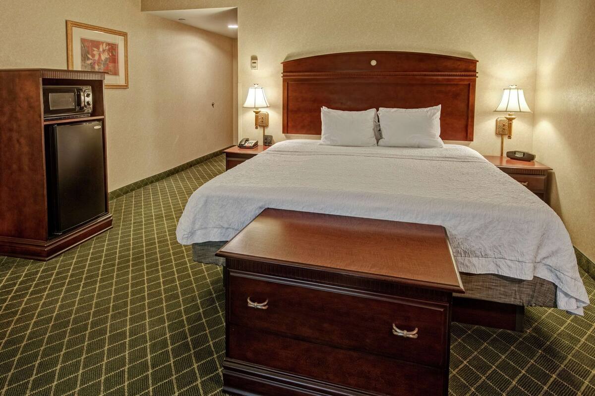 Habitación de hotel con una cama grande, lámparas de noche, una mesita de noche de madera, un refrigerador pequeño y un escritorio con cajones.