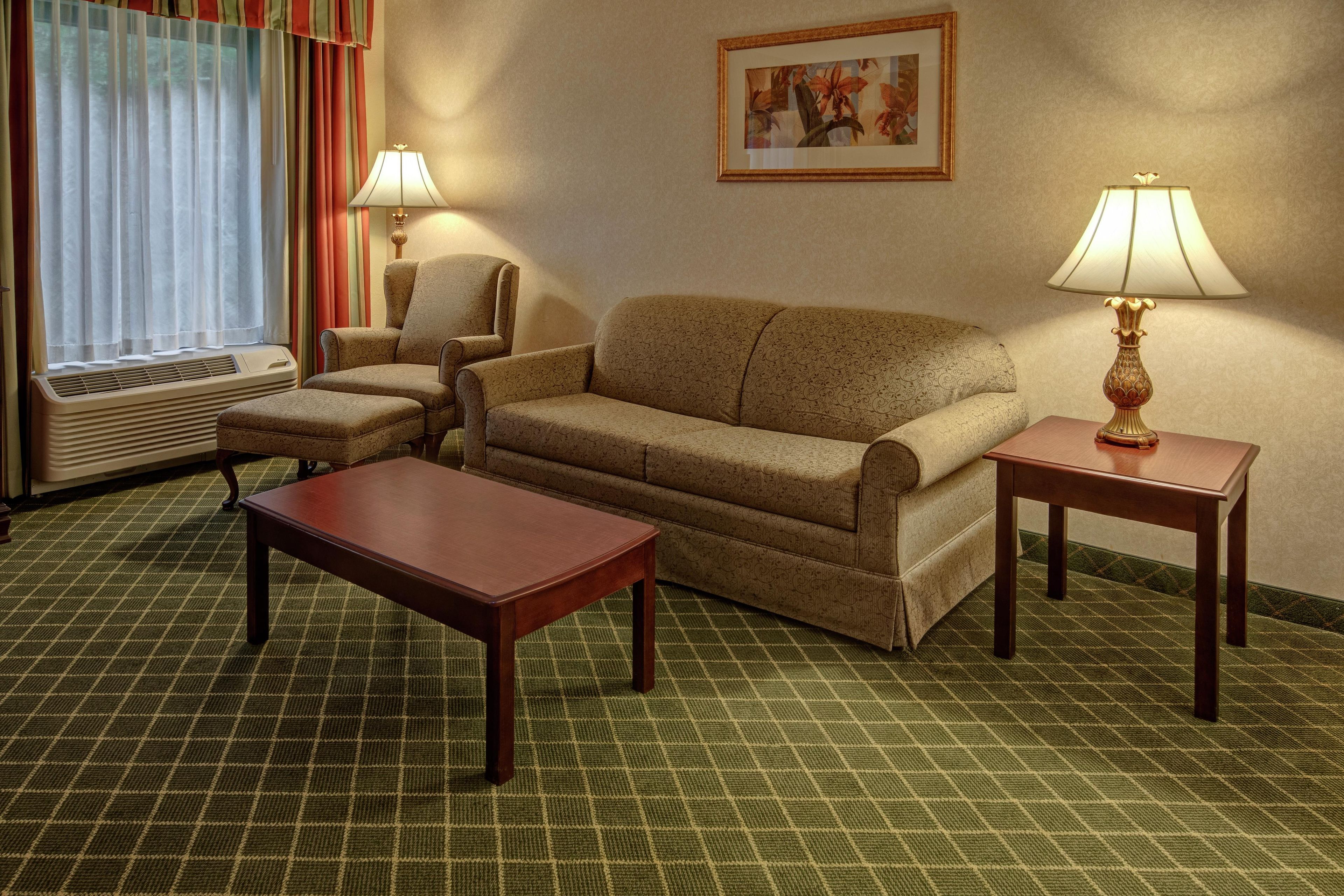 A hotel room with a sofa, two armchairs, a coffee table, two lamps, a framed picture on the wall, and an air conditioning unit.