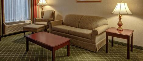 A hotel room with a sofa, two armchairs, a coffee table, two lamps, a framed picture on the wall, and an air conditioning unit.