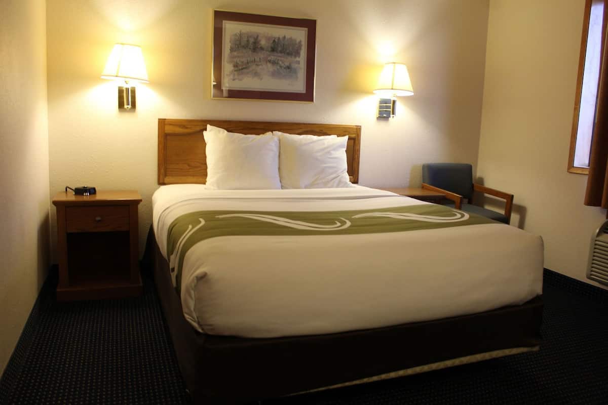 Quarto de hotel com cama, duas luminárias de cabeceira, mesa de cabeceira, cadeira e um quadro na parede.