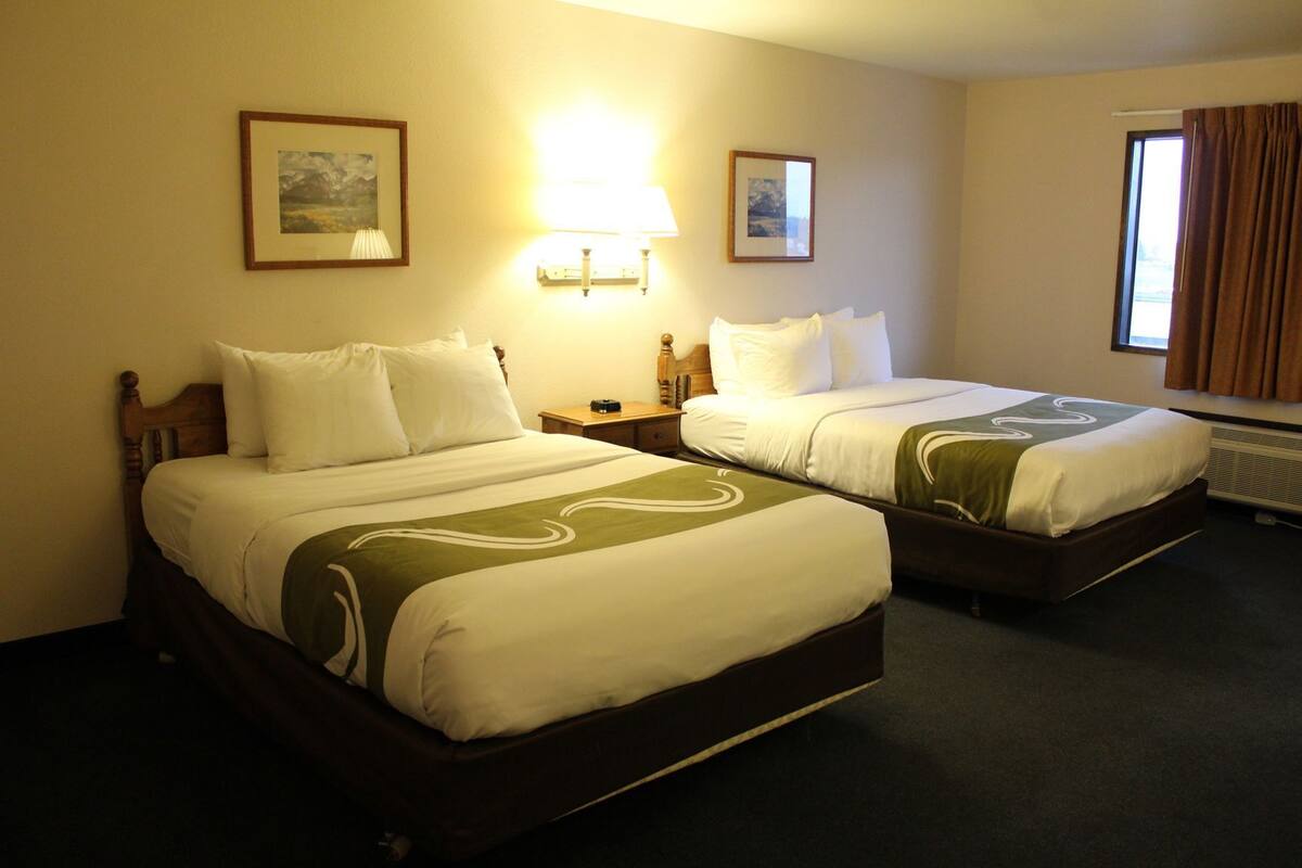 Quarto de hotel com duas camas, cada uma com roupa de cama branca e detalhes verdes. Há luminárias fixadas na parede e quadros emoldurados.