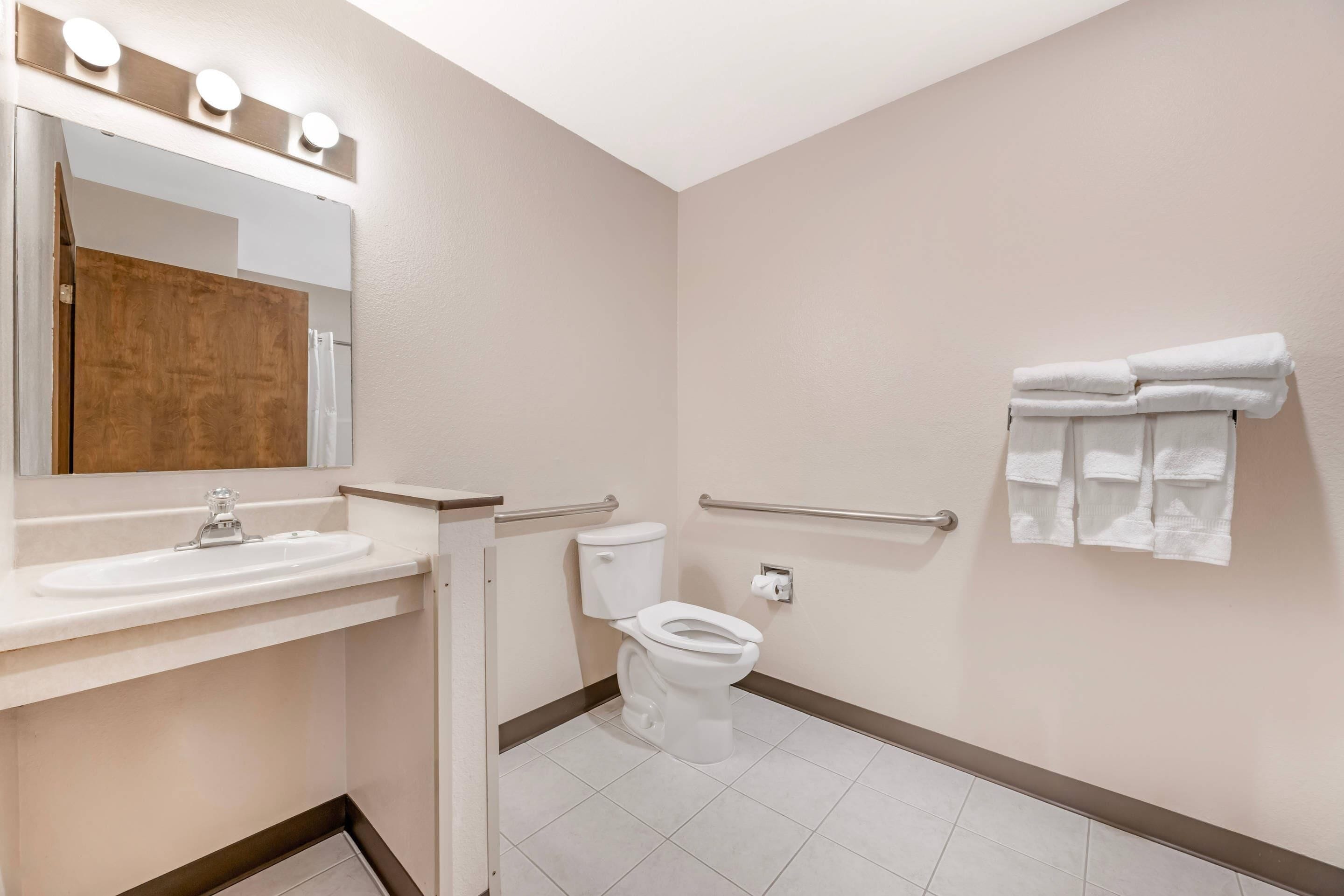 Un baño con lavamanos, espejo, inodoro y barras de apoyo.