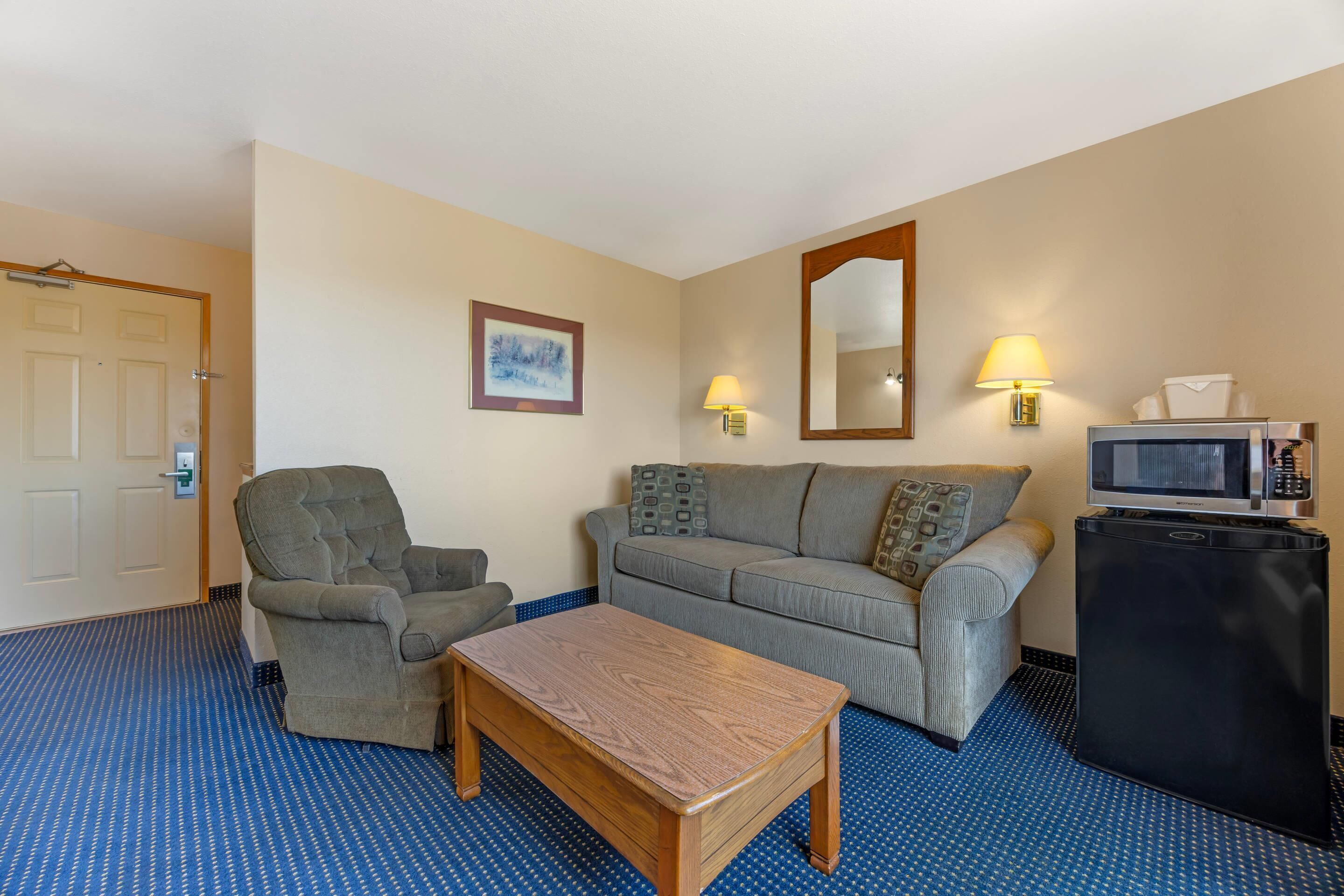 A hotel room with a sofa, armchair, wooden table, and a small refrigerator.