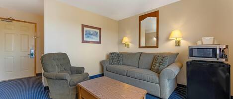A hotel room with a sofa, armchair, wooden table, and a small refrigerator.