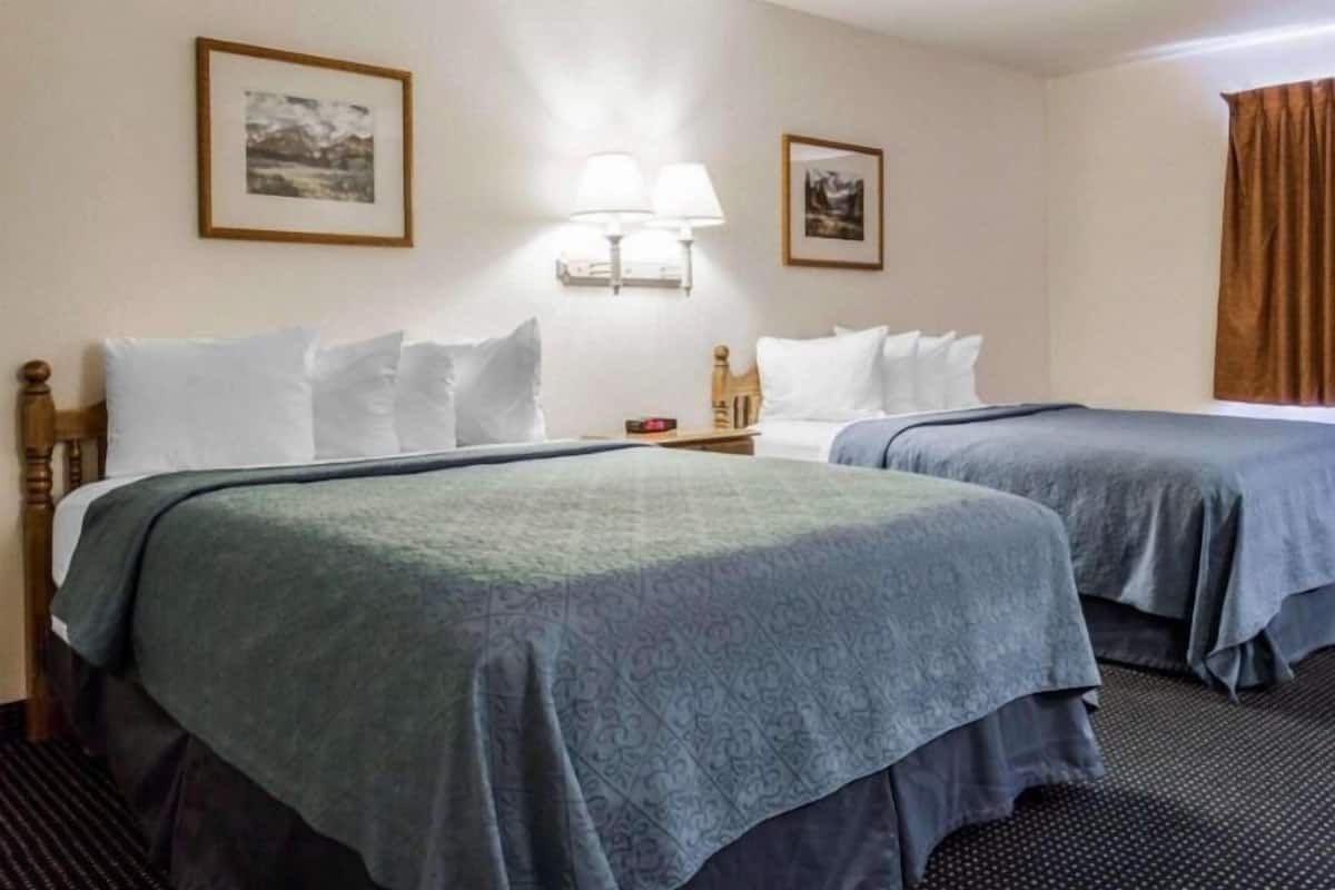 Quarto de hotel com duas camas, mesa de cabeceira, luminárias fixadas na parede e quadros emoldurados.