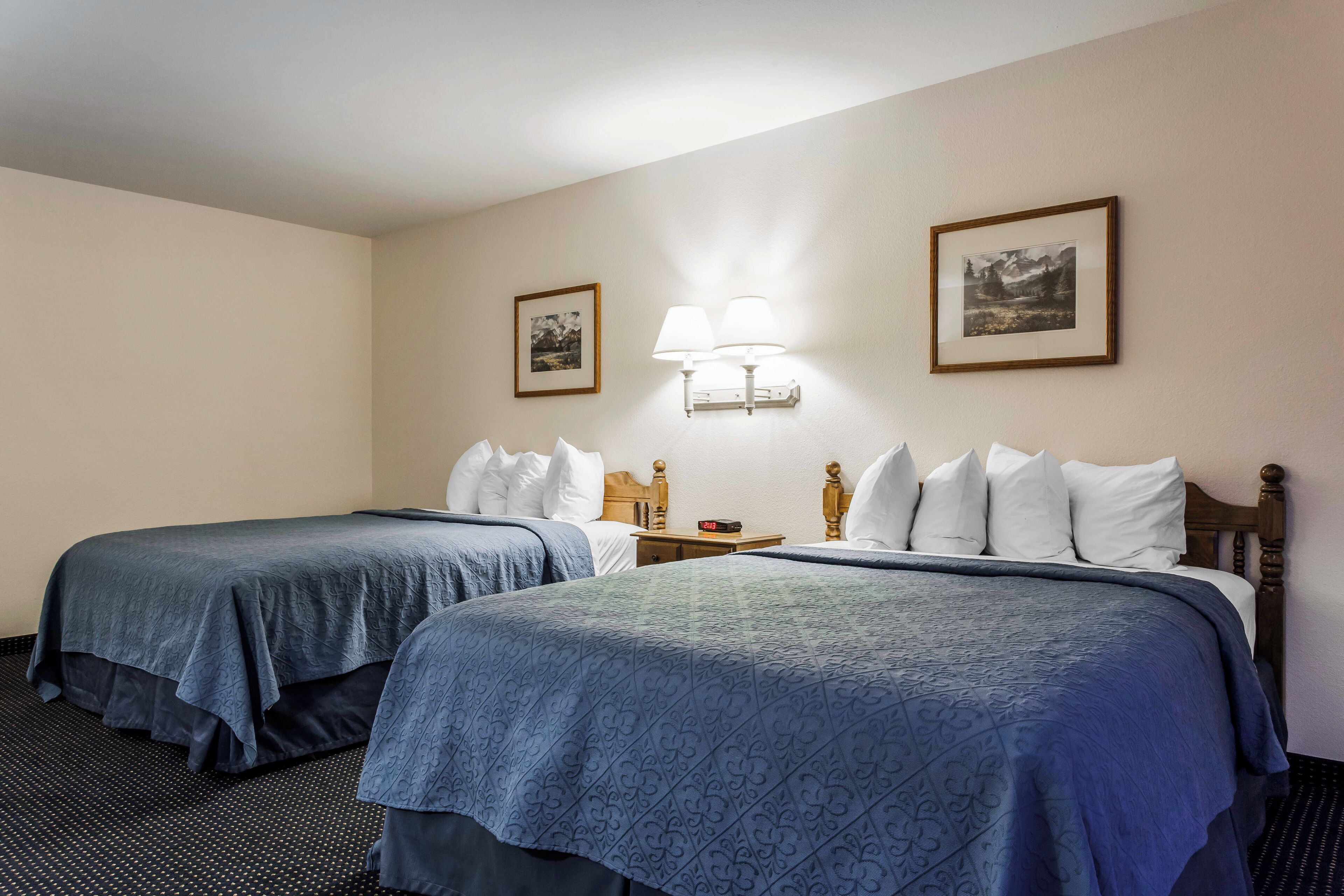 A hotel room with two beds, each with a blue bedspread and white pillows. There is a nightstand with a lamp between the beds.