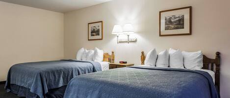 A hotel room with two beds, each with a blue bedspread and white pillows. There is a nightstand with a lamp between the beds.