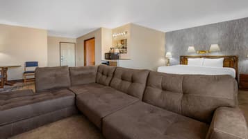 A hotel room with a sectional sofa, a bed with two pillows, a nightstand with lamps, and a kitchenette with a microwave.
