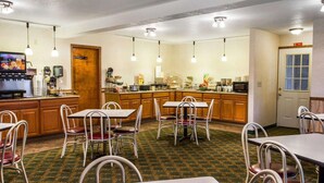 A hotel room with a kitchenette, dining area with tables and chairs, and a television displaying a menu.