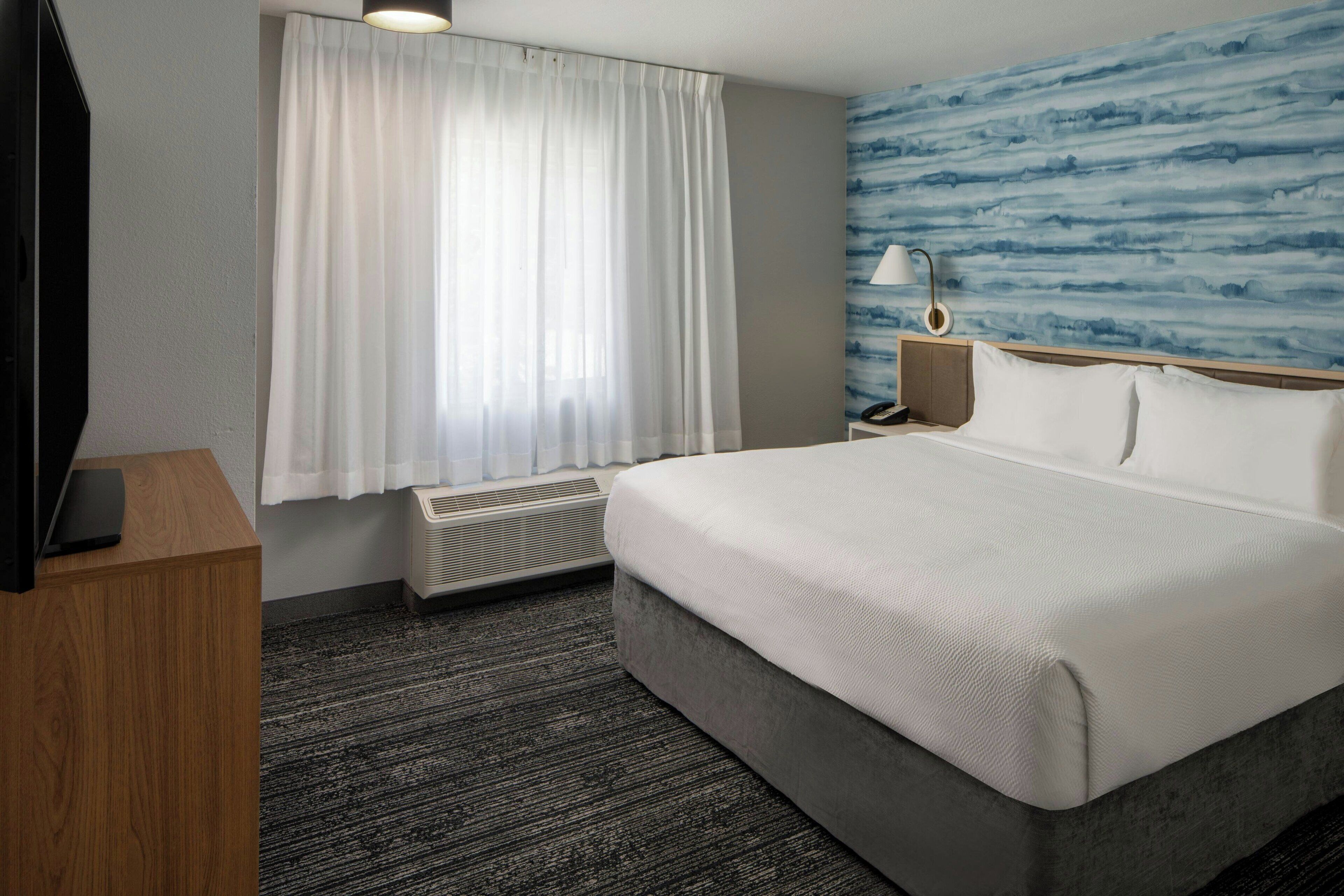 A hotel room with a large bed, a flat-screen TV on a wooden desk, a window with white curtains, a wall-mounted lamp, and a blue textured wall.