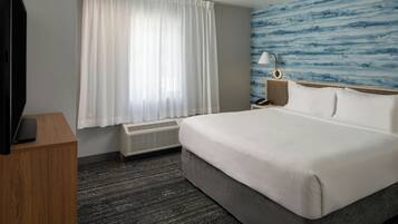A hotel room with a large bed, a flat-screen TV on a wooden desk, a window with white curtains, a wall-mounted lamp, and a blue textured wall.