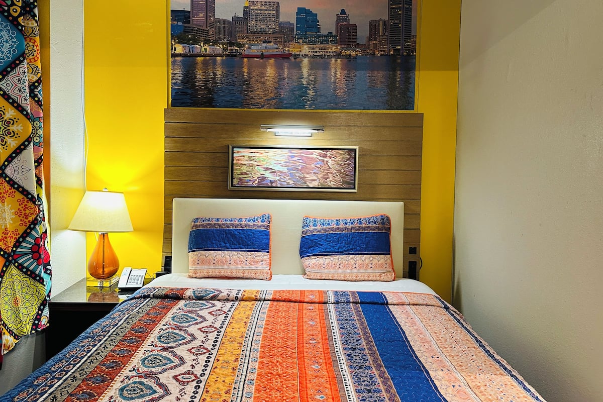 A hotel room with a bed, a nightstand, a lamp, a cityscape painting, and a patterned bedspread.
