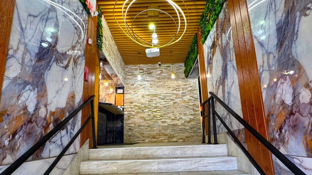 A marble staircase with a wooden finish, leading to a lobby area with a stone wall and a ceiling with circular light fixtures.