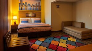 A hotel room with a bed, a sofa, a colorful rug, a cityscape painting, and a wooden nightstand with a lamp.